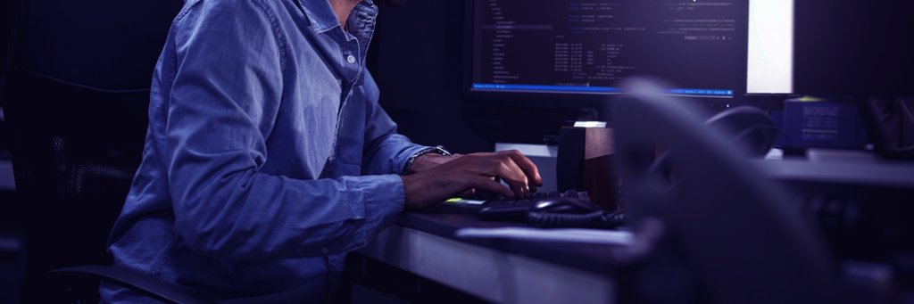 Profissional de tecnologia digitando em computador com telas de sistemas e códigos, representando monitoramento e gestão de infraestrutura de TI empresarial.