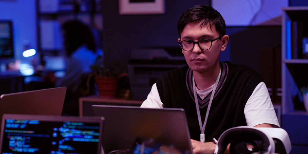 Profissional de tecnologia trabalhando em notebook em ambiente corporativo com códigos em tela, representando operação e gestão de sistemas de TI.